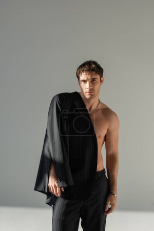 front view of brunette shirtless man in black trousers posing with stylish blazer on grey background