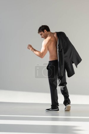 back view of shirtless man in black trousers and sneakers posing with blazer on grey background
