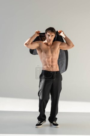full length of sexy muscular man in black pants and sneakers holding blazer and looking at camera on grey background