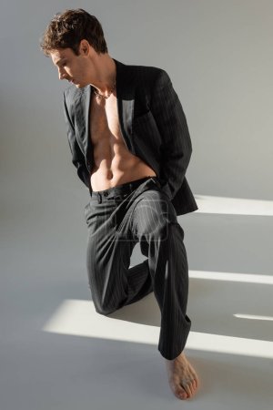 stylish barefoot man in black trousers and blazer on shirtless body posing on knee on grey background