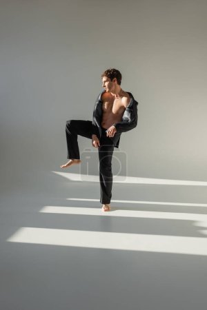 Téléchargez les photos : Full length of barefoot man in black trousers and blazer on shirtless body standing on one leg on grey background - en image libre de droit