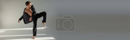 full length of trendy barefoot man in black pants and blazer on shirtless body posing on one leg on grey background, banner