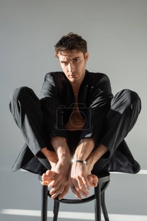 sexy barefoot man in black suit and silver bracelet sitting on chair and looking at camera on grey background