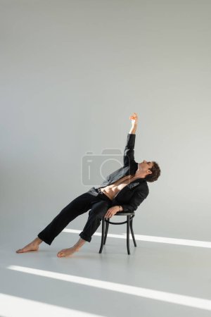 full length of barefoot man in black suit posing on chair with raised hand on grey background with copy space
