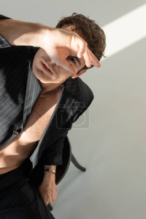 Photo for Top view of man wearing black blazer on shirtless body obscuring face with hand and looking at camera on grey background - Royalty Free Image