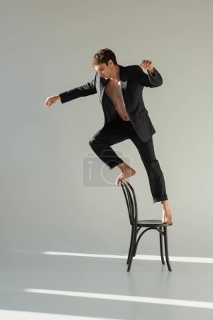 full length of barefoot and shirtless man in black suit doing trick while standing on chair on grey background