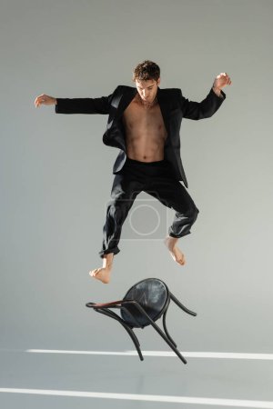 full length of muscular man in black stylish blazer jumping from chair on grey background