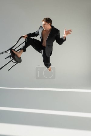 full length of barefoot man in trendy suit jumping with chair on grey background with lighting