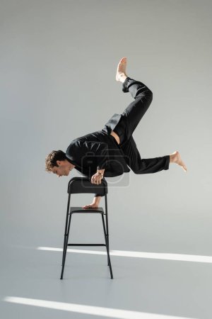 full length of trendy barefoot man doing handstand on chair on grey background