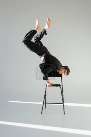 full length of barefoot man in black suit doing handstand on chair and looking at camera on grey background