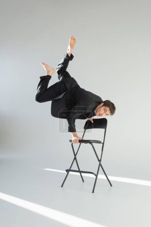 full length of barefoot man in black trendy suit looking at camera and doing handstand on chair on grey background