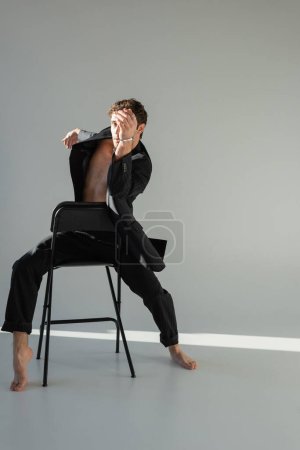 full length of trendy barefoot man in black suit sitting on chair and covering face with hand on grey background