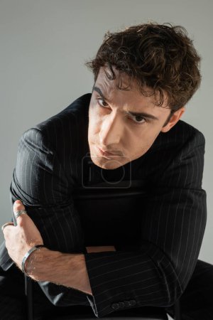 brunette men in black blazer and metal bracelet sitting with crossed arms and looking at camera isolated on grey