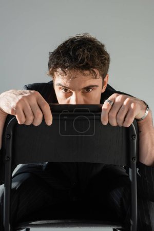 brunette man in black clothes hiding face behind chair back and looking at camera isolated on grey