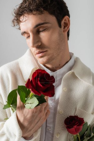 stylish man in white soft jacket standing with closed eyes and holding red roses isolated on grey