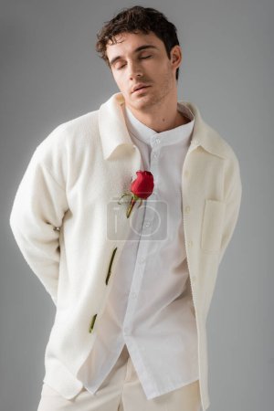 trendy man in white jacket decorated with red roses standing with closed eyes isolated on grey