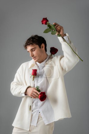 Téléchargez les photos : Man in white stylish clothes posing with red roses and closed eyes isolated on grey - en image libre de droit