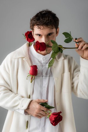 fashionable man in white soft jacket obscuring face with red rose while looking at camera isolated on grey Poster 634762380