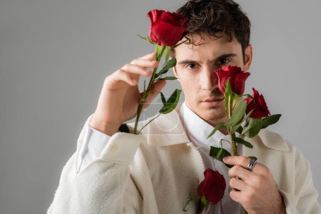 Foto de Stylish man in white jacket and silver finger ring holding red roses and looking at camera isolated on grey - Imagen libre de derechos