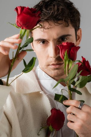 Foto de Portrait of man in white jacket and silver finger ring looking at camera and holding red roses isolated on grey - Imagen libre de derechos