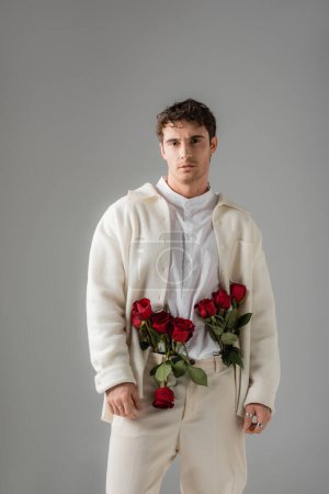 front view of man in white stylish clothes standing with red roses in pants isolated on grey