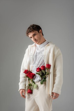 stylish man looking at camera while posing with red roses in white trousers isolated on grey
