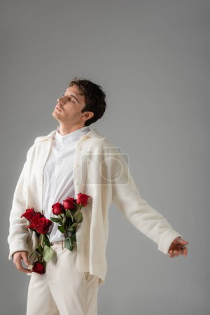 stylish man in white clothing posing with closed eyes and red roses in trousers isolated on grey