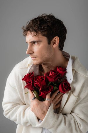 brunette man in white soft jacket and silver finger rings holding bouquet of red roses and looking away isolated on grey