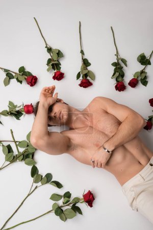 top view of sexy shirtless man in silver bracelet lying near red roses on white background
