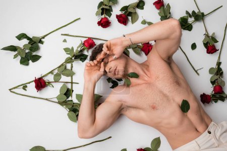 top view of shirtless muscular man with closed eyes lying near red roses on white background