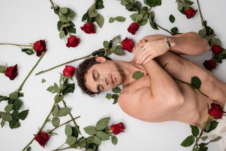 top view of shirtless man in silver bracelet and finger ring lying with closed eyes near red roses on white background