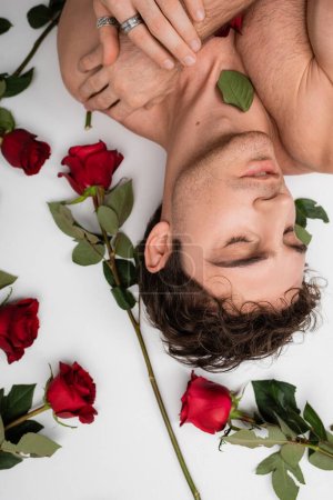 top view of shirtless man in silver finger rings lying with closed eyes near red fresh roses on white background