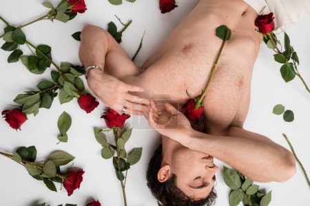 top view of sexy man with shirtless torso and closed eyes lying near red roses on white background