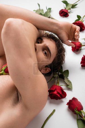 high angle view of sexy shirtless man obscuring face and looking at camera while lying near red roses on white background