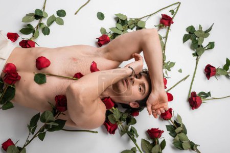 Foto de Top view of sexy shirtless man lying with hands near face and looking at camera near red roses on white background - Imagen libre de derechos