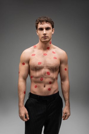 front view of sexy shirtless man with red kiss prints on body looking at camera on grey background