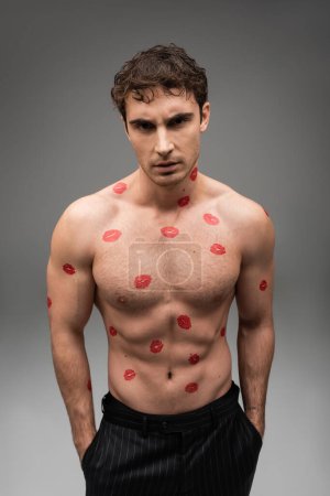 shirtless man with red kisses on muscular body standing with hands in pockets of black pants on grey background