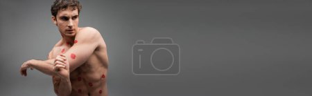 sexy brunette man with red kiss prints on muscular body looking away on grey background, banner