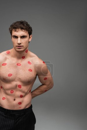 Photo for Brunette man with red lipstick prints on shirtless muscular torso looking at camera on grey background - Royalty Free Image