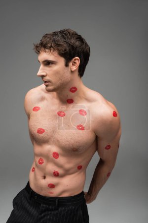 brunette man with red kiss prints on shirtless torso looking away on grey background