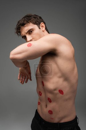 Photo for Sexy shirtless man with red kiss prints on body obscuring face with arm while looking at camera isolated on grey - Royalty Free Image