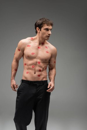 Photo for Sexy man in black pants posing with red kiss prints on shirtless muscular torso and looking away on grey background - Royalty Free Image