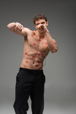 athletic man with red kisses on shirtless body obscuring face while posing with closed eyes on grey background
