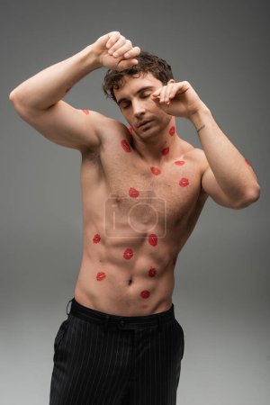 sexy shirtless man with red lipstick marks on body posing with closed eyes on grey background