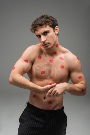 athletic man with red lip prints on shirtless muscular body looking at camera on grey background