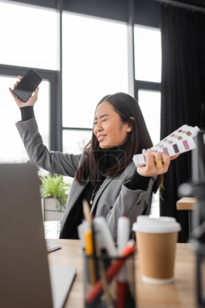 Excited asian interior designer holding smartphone and color samples in studio 