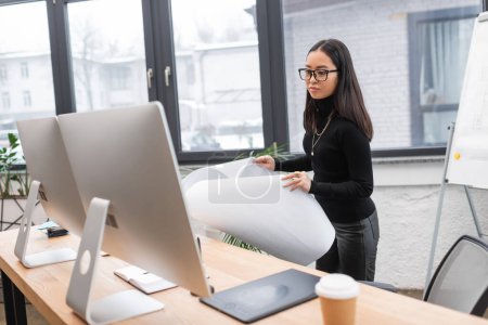 Asian interior designer holding blueprint near computers in office 