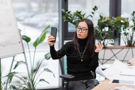 Brunette asian designer in eyeglasses having video call on smartphone near blueprints in office 