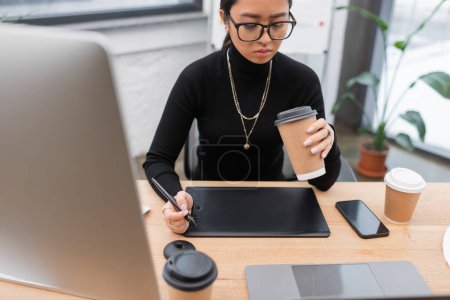 Asian designer looking at paper cup near gadgets and graphic tablet in office 