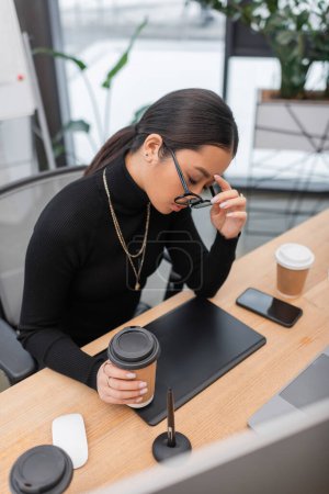 Exhausted asian designer in eyeglasses holding paper cup near gadgets in studio 